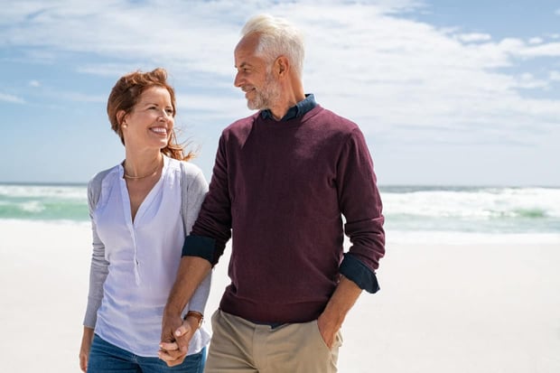 Happy senior couple walking on the beach in a sunny day. Smiling mature couple looking at each other on beach during sunset with copy space. Retired man in love with his wife relaxing during vacation.