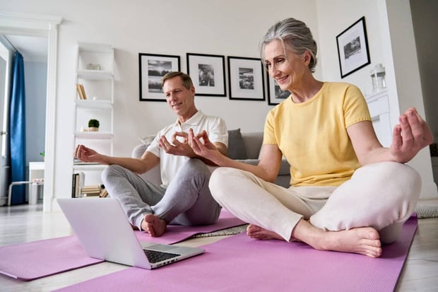 Happy old middle aged senior 50s couple learning to meditate at home watching live online tv yoga class tutorial on website looking at laptop computer doing virtual training fitness workout exercises.