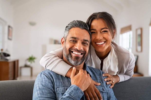 Portrait of multiethnic couple embracing and looking at camera sitting on sofa. Smiling african american woman hugging mid adult man sitting on couch from behind at home. Happy mixed race couple laugh Portrait of multiethnic couple embracing and looking at camera sitting on sofa. Smiling african american woman hugging mid adult man sitting on couch from behind at home. Happy mixed race couple laugh