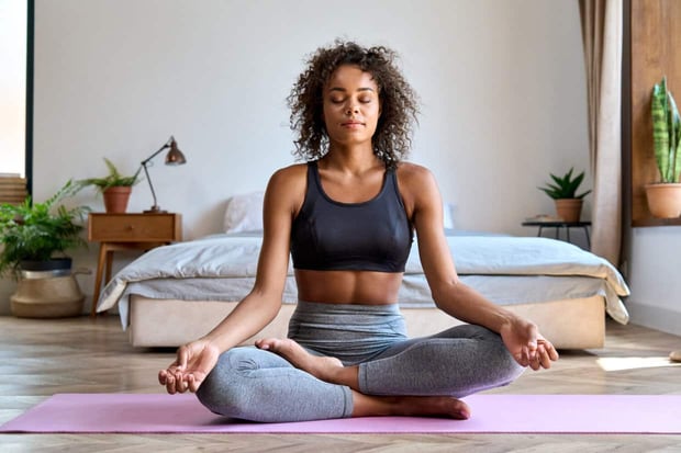 Young calm fit healthy African American woman wearing sportswear sitting at home in bedroom doing yoga exercise, meditating and breathing in the morning. Mental health and zen meditation concept