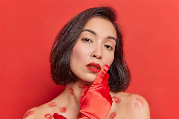 Portrait of gentle young Asian woman with clean healthy skin looks tenderly at camera has natural dark hair poses for magazine cover looks seductive isolated over vivid red studio background Portrait of gentle young Asian woman with clean healthy skin looks tenderly at camera has natural dark hair poses for magazine cover looks seductive isolated over vivid red studio background
