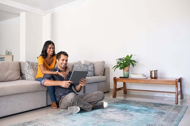 Young middle eastern couple using digital tablet while sitting on couch and floor. Happy smiling indian woman embracing from behind her boyfriend while watching video on digital tablet at home.