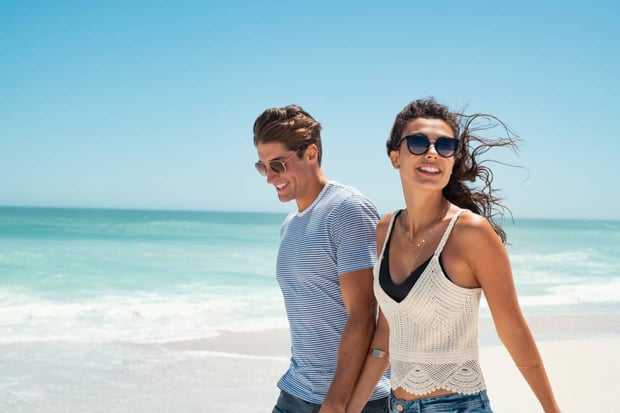 Beautiful woman with man wearing sunglasses walking on beach.. Young couple enjoying honeymoon after marriage at sea. Happy casual couple holding hands and walking at the beach with copy space. Beautiful woman with man wearing sunglasses walking on beach.. Young couple enjoying honeymoon after marriage at sea. Happy casual couple holding hands and walking at the beach with copy space.