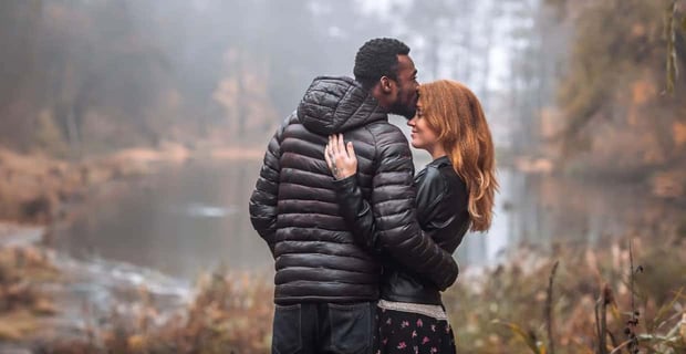 Photo of a Black man kissing a red haired woman on the head as they cuddle in a forest Photo of a Black man kissing a red haired woman on the head as they cuddle in a forest