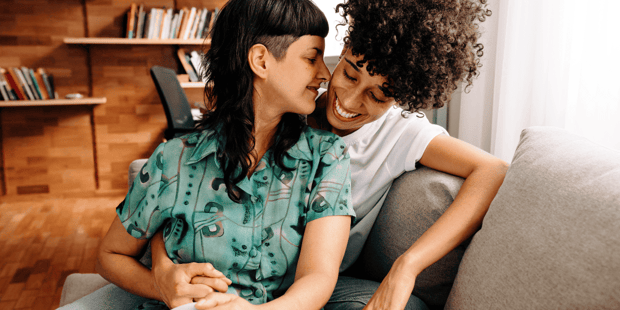Queer interracial couple sitting together on couch, smiling and almost kissing