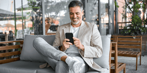 Handsome older man sitting in suit on couch with leg on knee scrolling on phone
