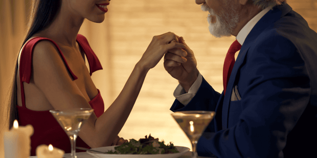 Older man and younger woman in red dress at a fancy dinner with man holding young woman's hand