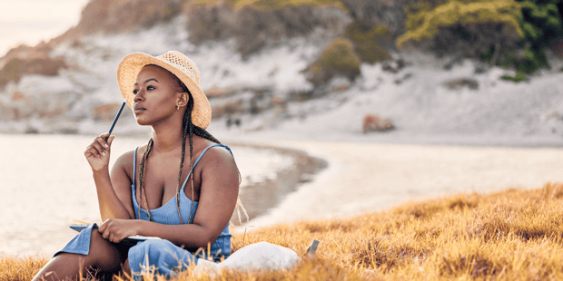 Black woman sitting on beach with a notebook pondering