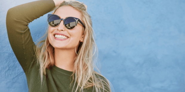 Young blonde woman in olive green shirt and sunglasses smiling wide and holding hair