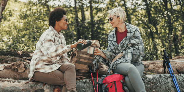 A Black and a white woman couple pouring coffee for each other while camping or hiking