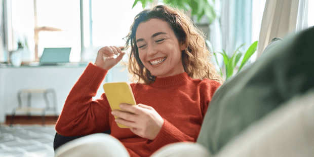 Young woman with curly hair lounging on couch and laughing at cellphone