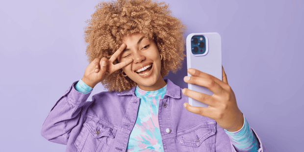 Woman with blonde curly hair holding up peace sign to take a selfie in front of a purple wall
