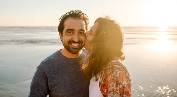 Photo of a woman kissing a man's cheek at the beach while the man smiles at the camera
