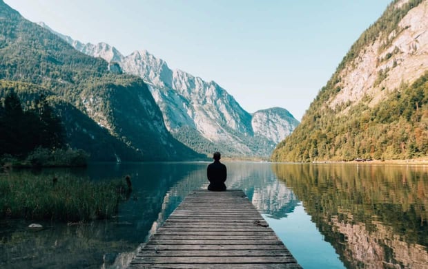 Photo of a man sitting alone on a pier overlooking a lake and mountainside