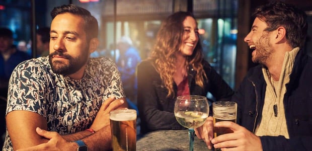 Man looking away dejected while two people flirt at a bar